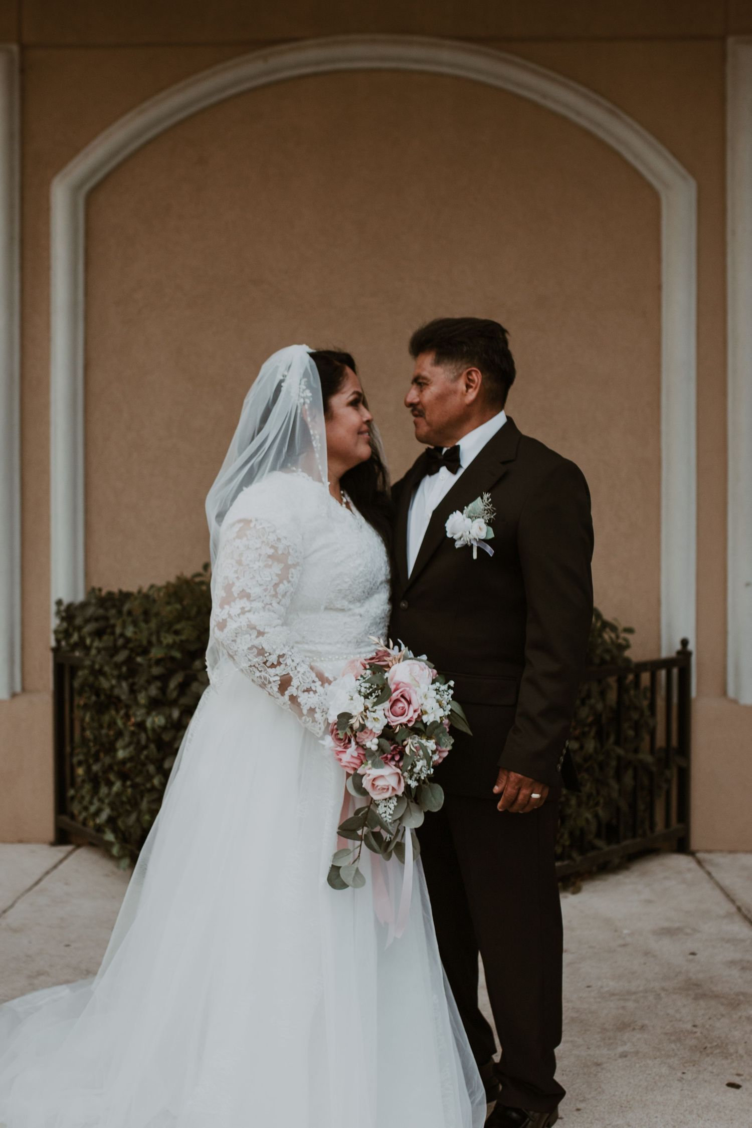 Bride and groom in formal wedding attire standing before an arched doorway, bride holding pink bouquet.