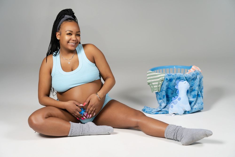 Person in athletic wear smiles while sitting on floor with blue bag against gray studio background.