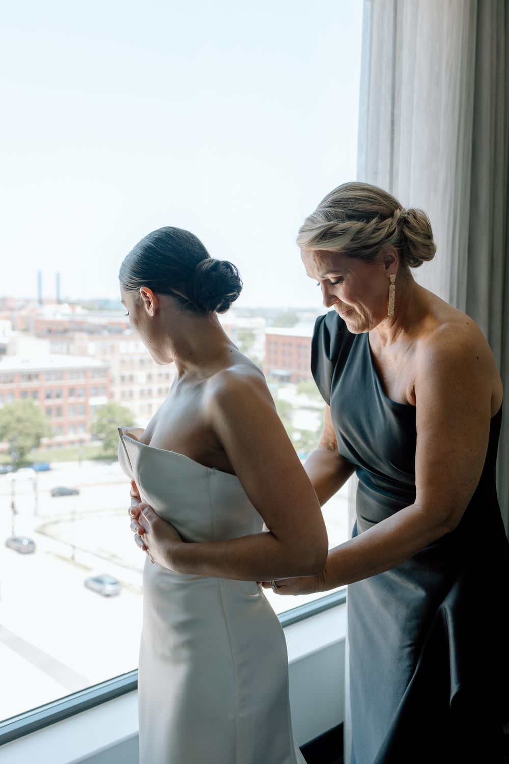 Bridesmaids in gray dresses help with wedding preparations near a bright window.