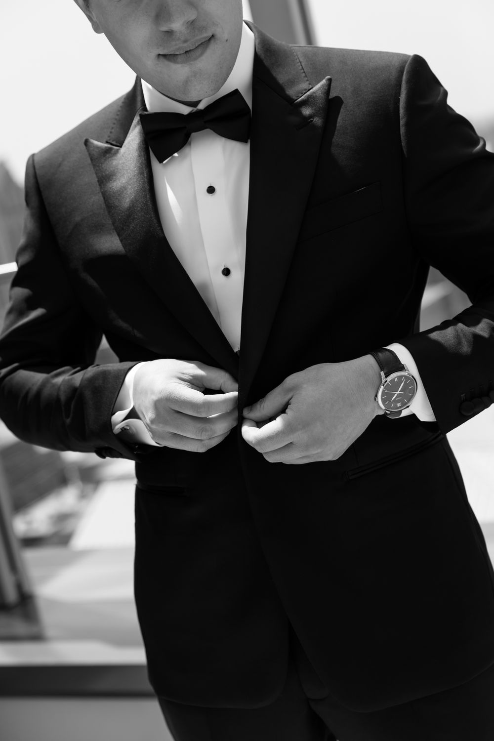 Groomsmen in black tuxedos and bow ties adjust their cuffs and buttons in a formal wedding preparation moment.