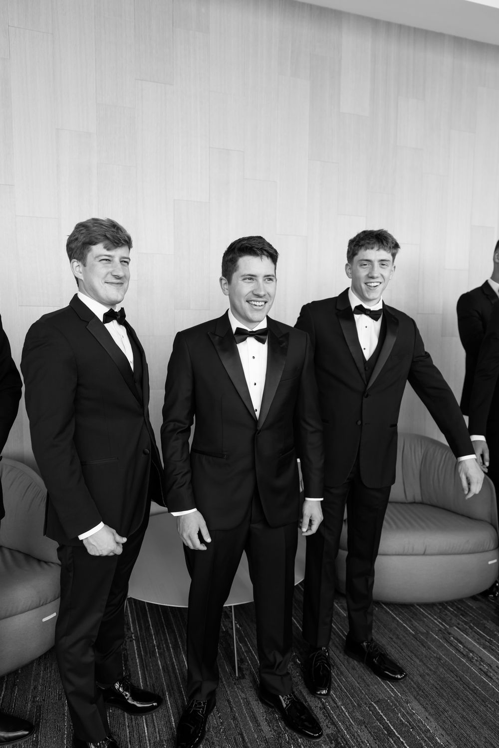 A group in formal black tuxedos sitting together and laughing in a casual moment.