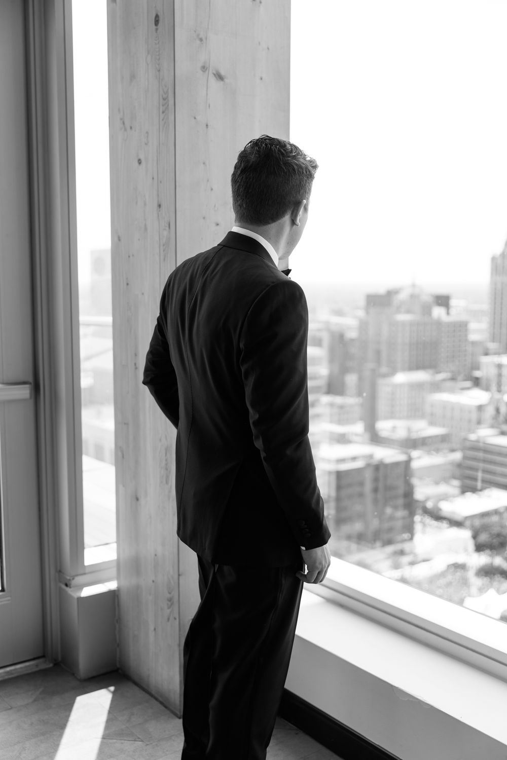 Black and white photo of a person in formal attire gazing out large windows at a city skyline view.