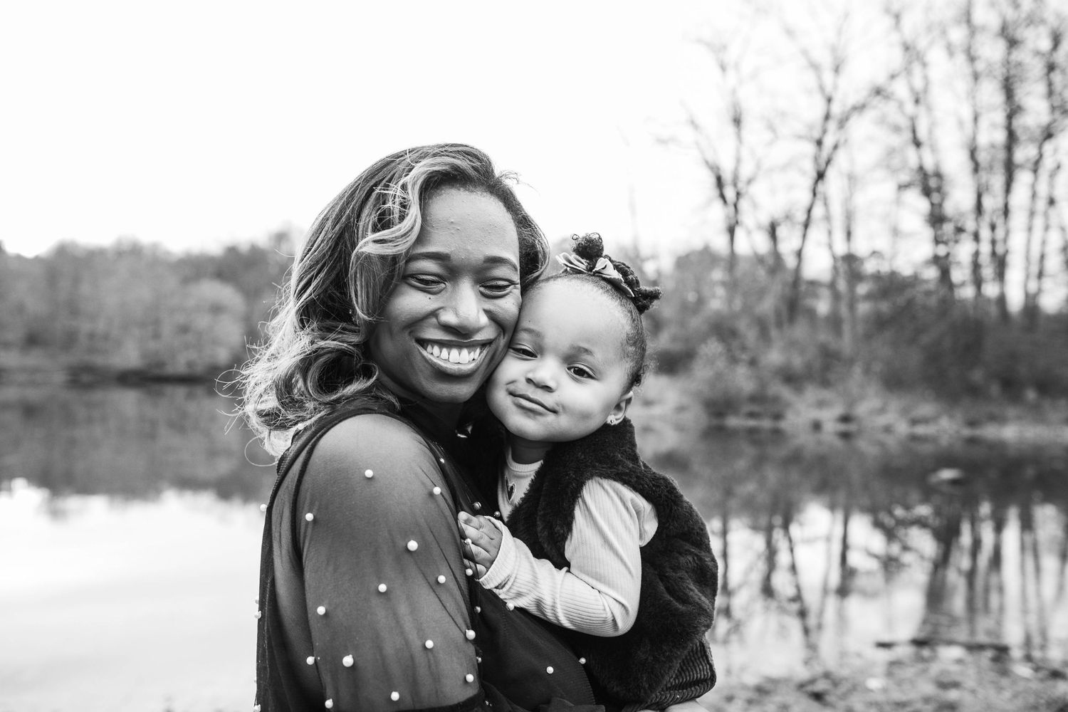 A smiling mother holds her happy baby girl in a black and white outdoor portrait by a lake.