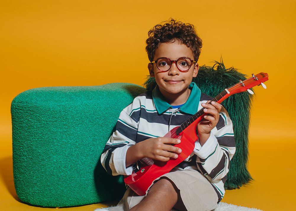 Person in glasses sits on green couch with red guitar against orange wall in creative photo series.