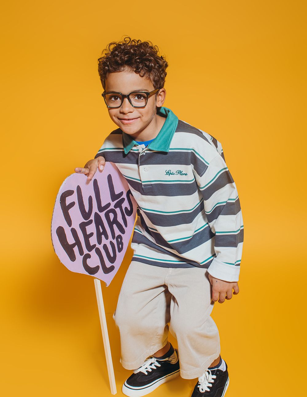 A child in a striped shirt and glasses poses with 'hello' signs against a vibrant orange background.