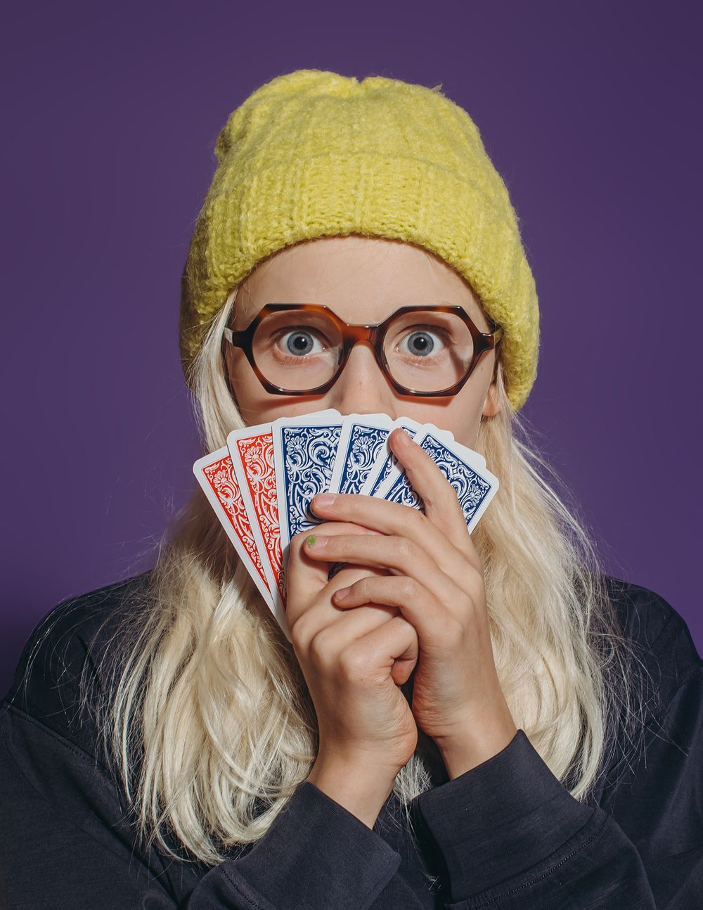 A person in glasses and yellow beanie holds playing cards in a sequence of four photos against a purple background.
