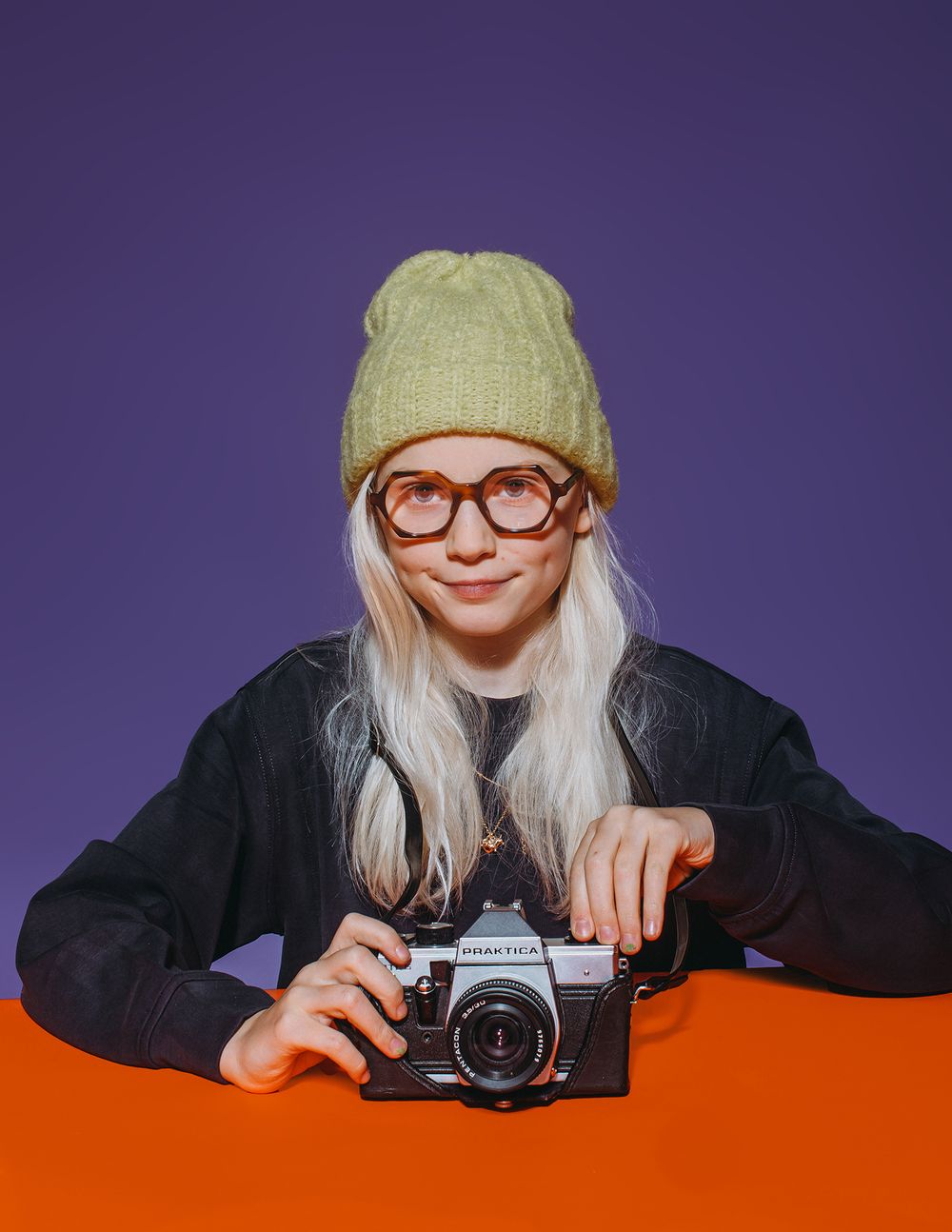 A person in a knit hat and red glasses peeks over an orange surface while holding a vintage camera.