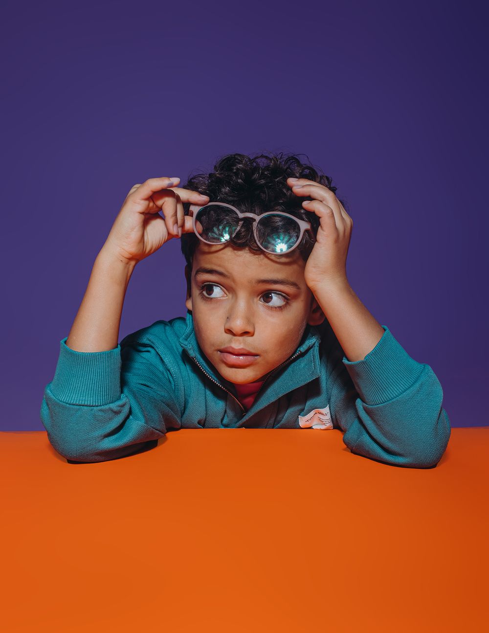 A person in a turquoise hoodie playfully peeks over an orange surface while adjusting sunglasses.