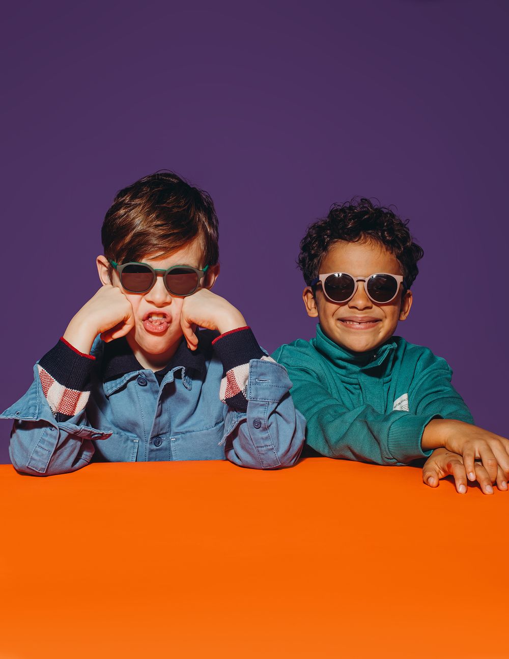 Group of children wearing sunglasses and denim jackets pose against purple and orange background in retro style photo.