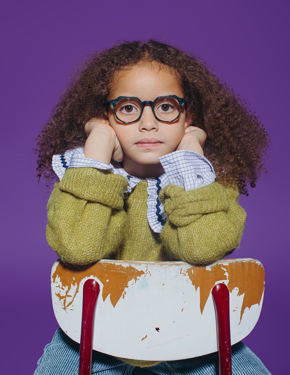 Person with curly hair and glasses rests head on hands while seated against purple backdrop.