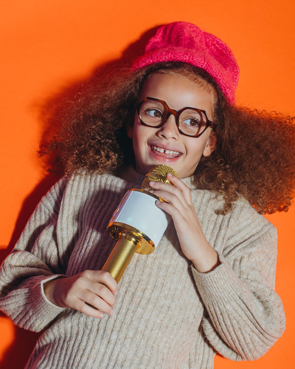 Person in pink beanie and glasses holding glass bottle against vibrant orange background in creative photo series.