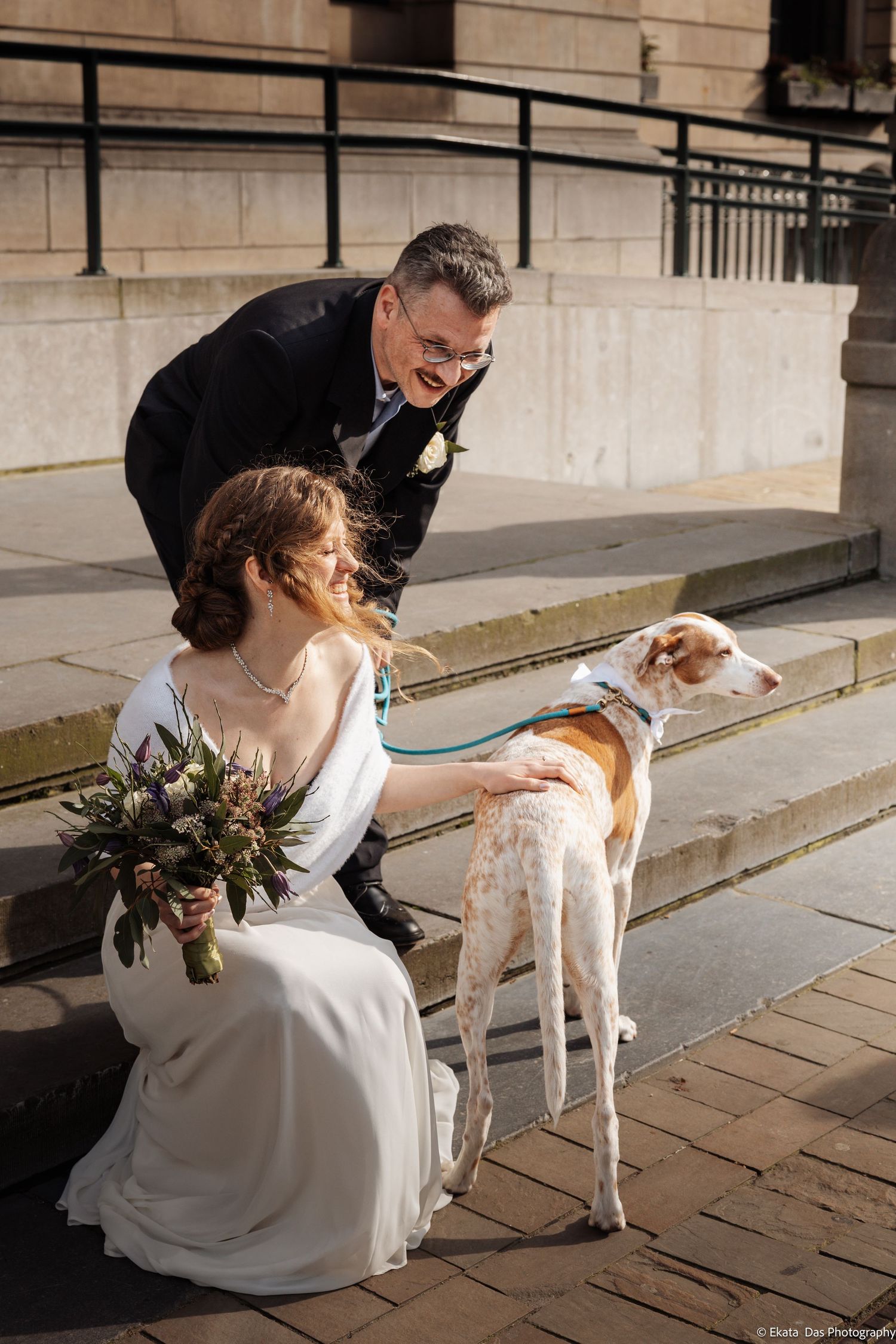 A bride in white dress pets a friendly white dog on stone steps during a wedding photoshoot.