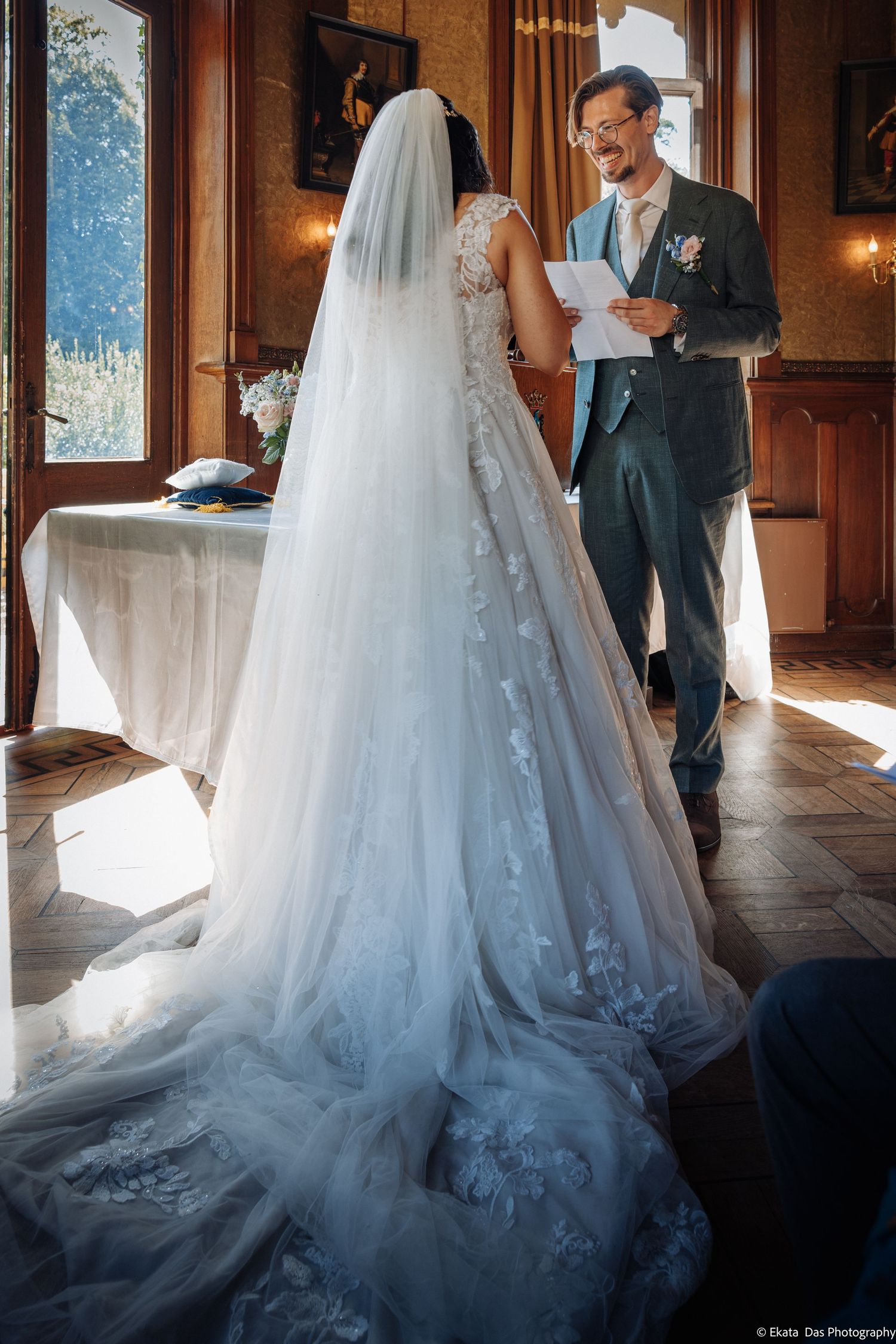 Bride in lace wedding gown with veil faces groom reading vows during indoor wedding ceremony.