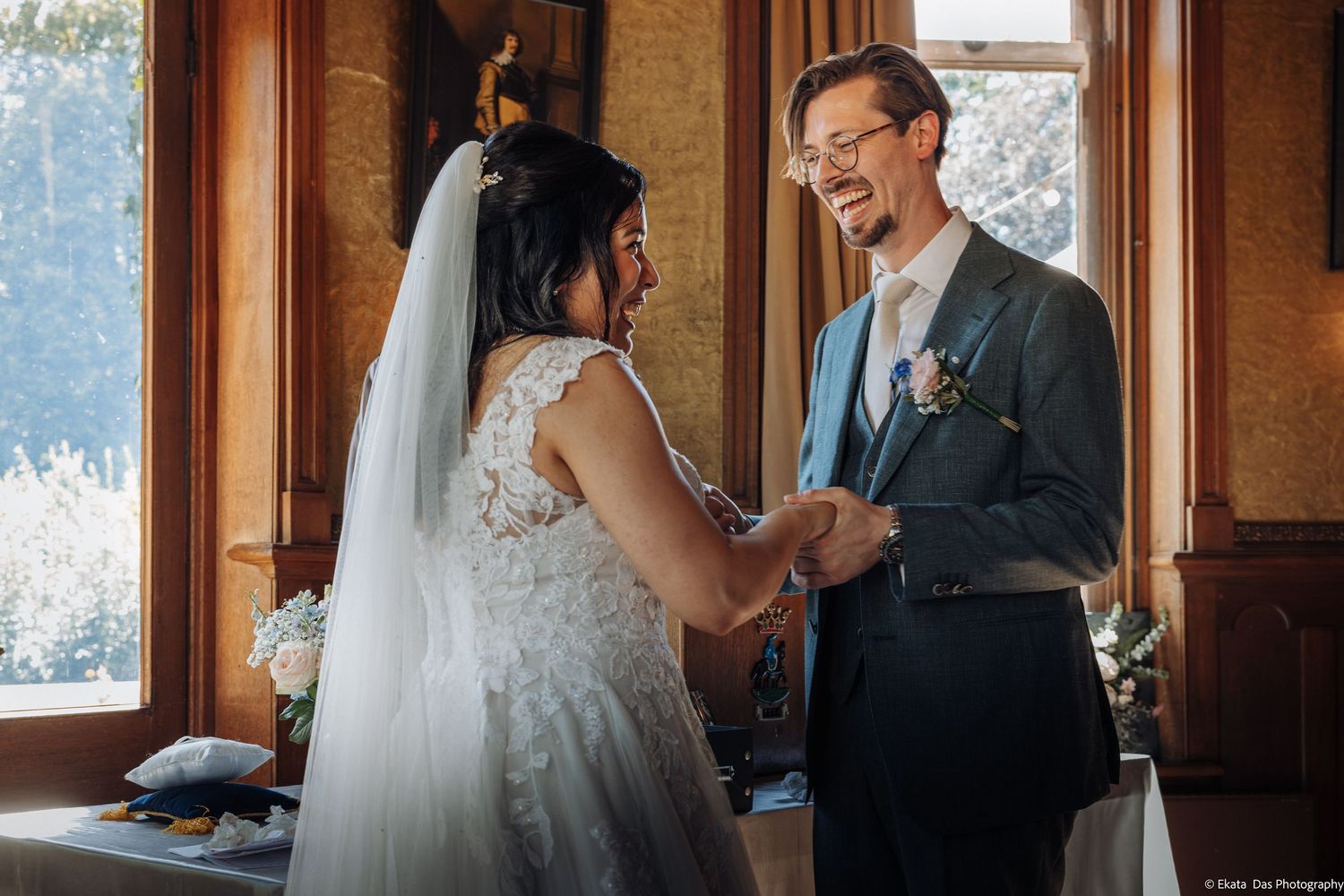 Bride and groom laughing together while exchanging vows indoors, groom in gray suit, bride in lace wedding dress with veil.