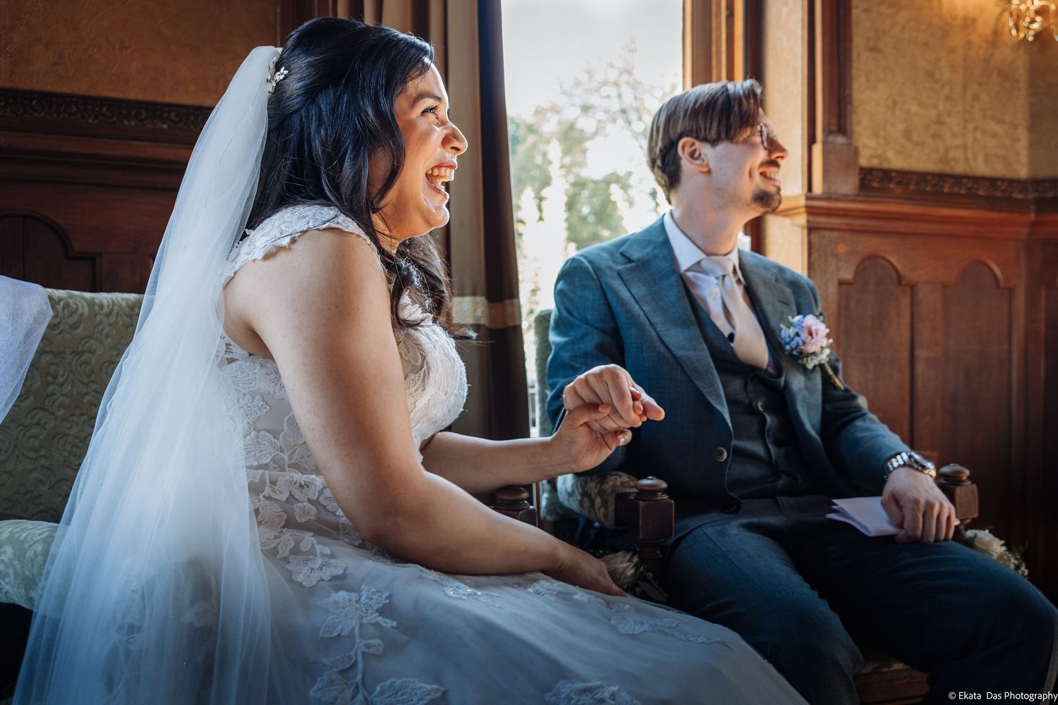 Bride and groom laughing joyfully during wedding ceremony, seated together in elegant venue with ornate decor.