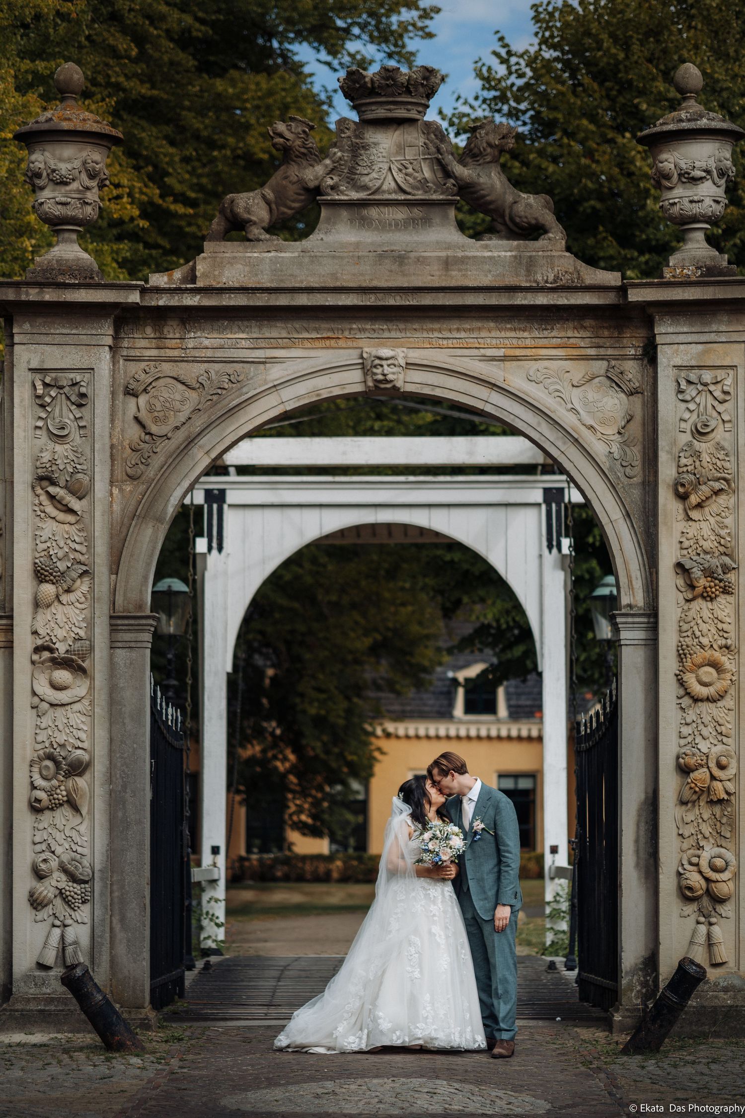 Bride and groom kissing under ornate stone archway with crown and lion sculptures at elegant wedding venue.
