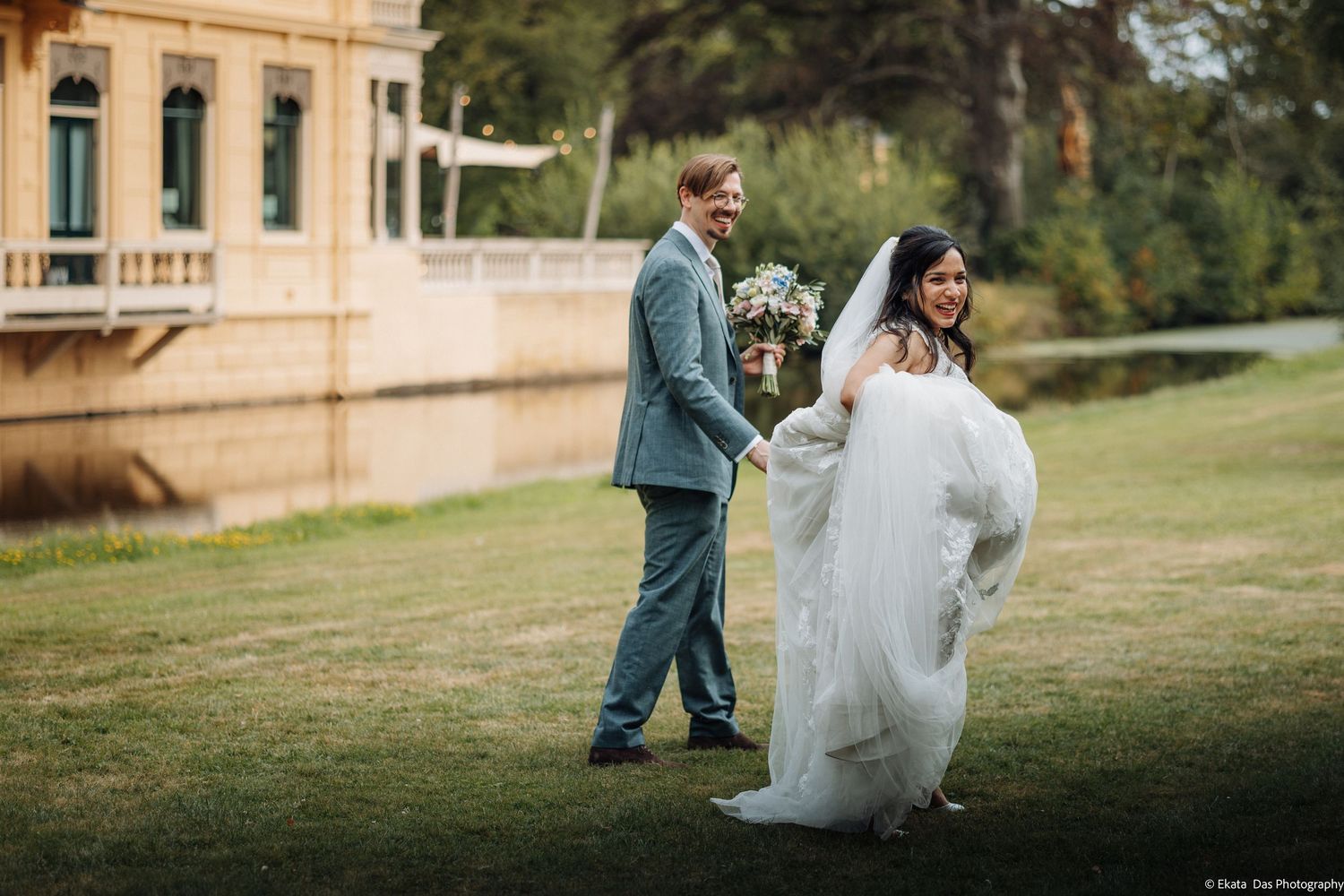 A bride in a flowing white gown and groom in gray suit walk hand in hand on lush green lawn near elegant venue.