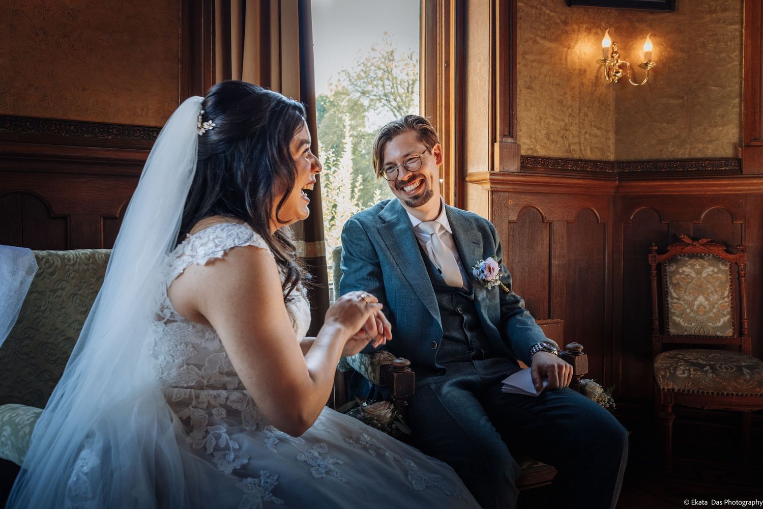 Happy bride and groom sharing a laughing moment together while seated on a sofa in a classic elegant venue.