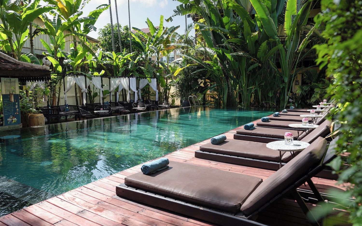 Luxurious pool loungers line the edge of a tranquil turquoise swimming pool surrounded by lush tropical gardens.