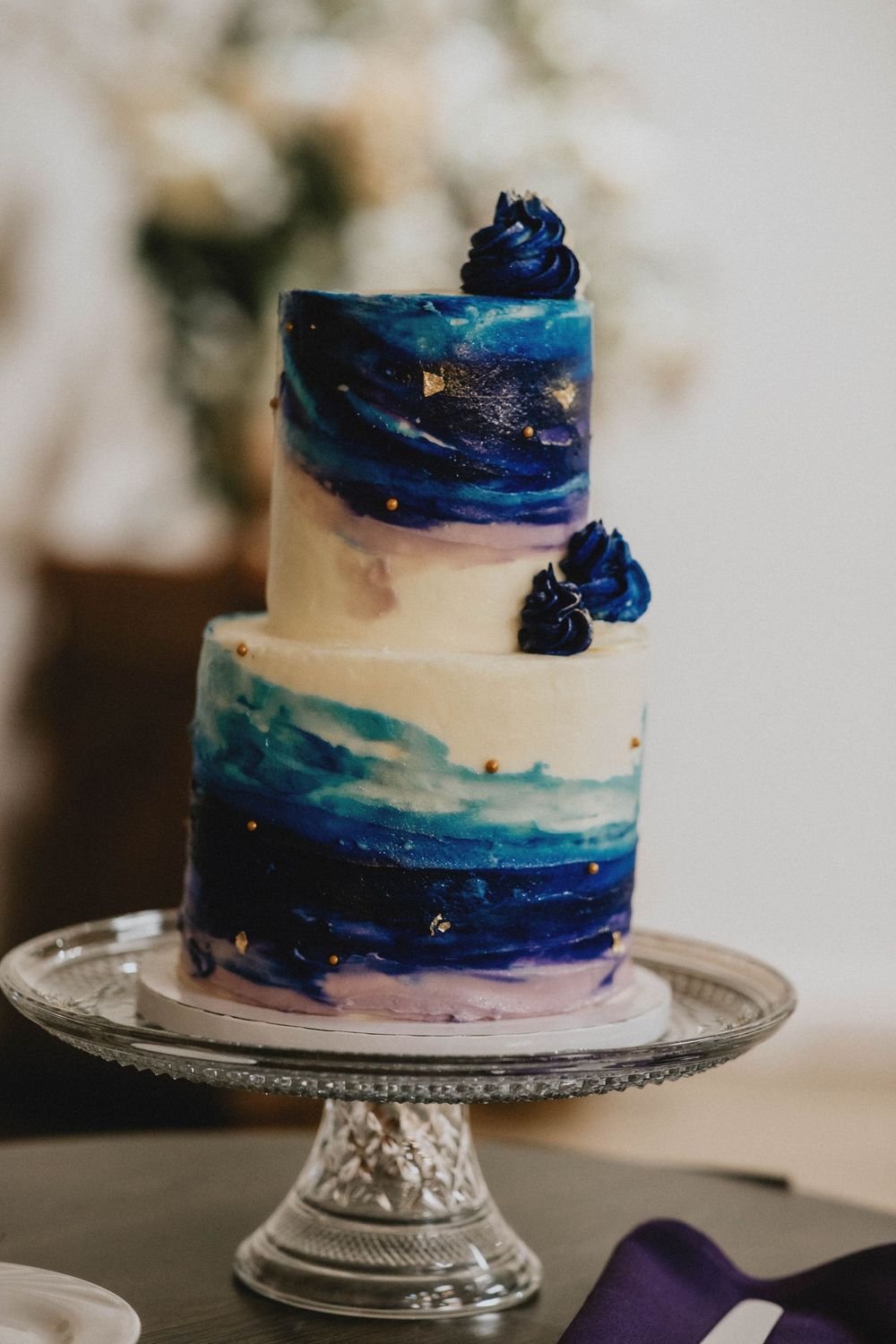 A white wedding cake decorated with navy blue and purple watercolor-style brushstrokes sits elegantly on an ornate cake stand.