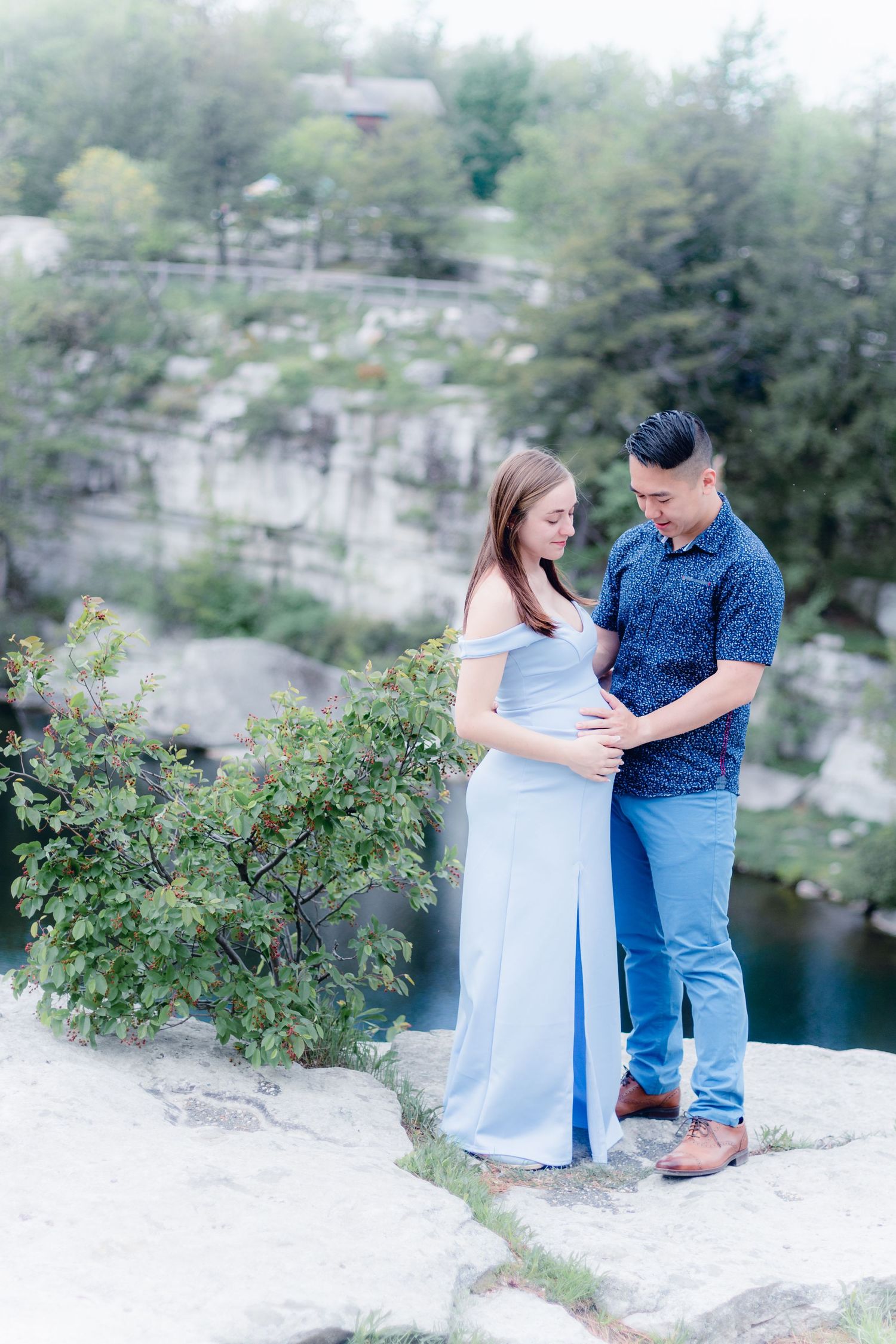 A couple shares a tender moment together on a rocky overlook near a serene body of water on a sunny day.