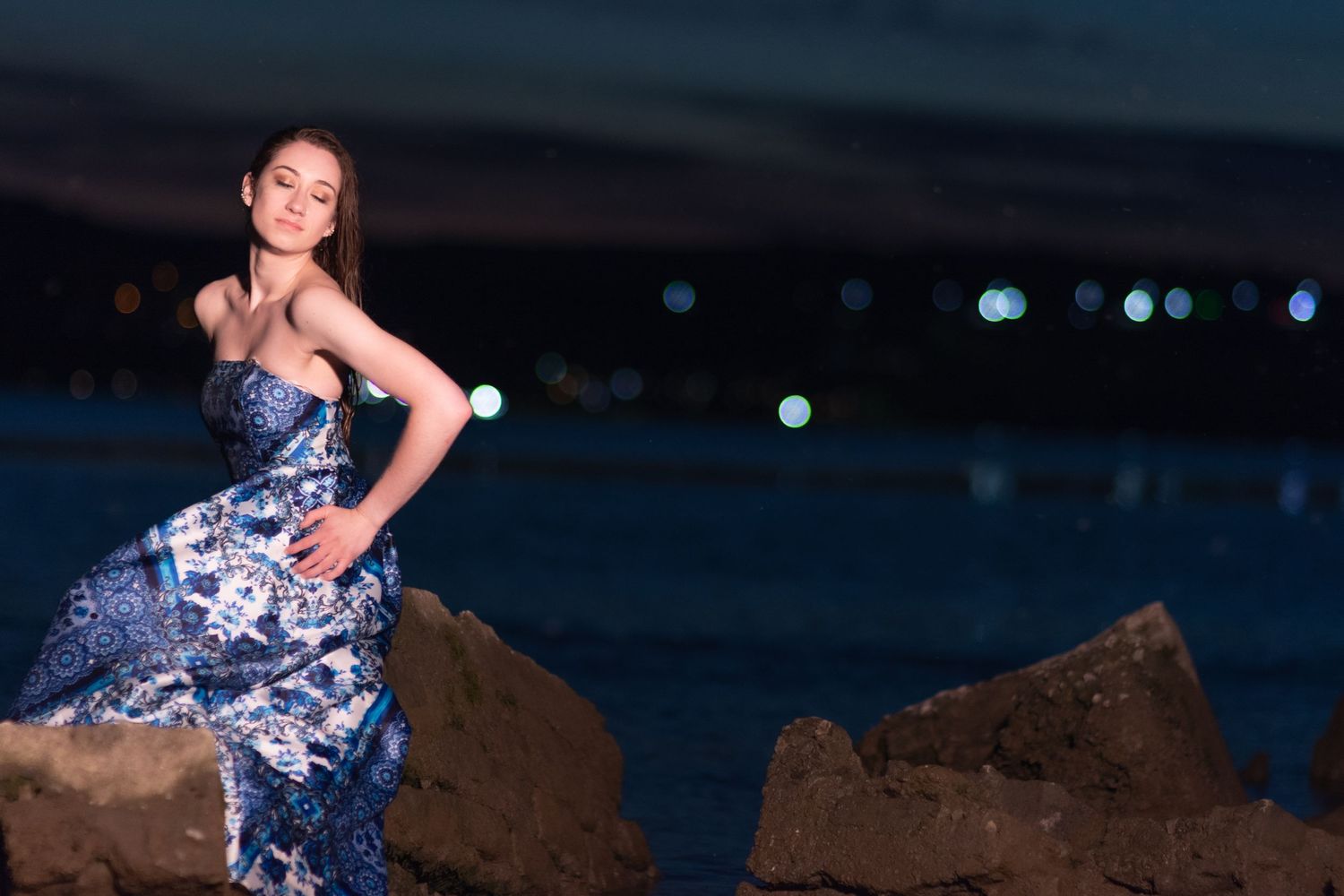 A figure in a blue floral dress poses gracefully against a dark evening sky with bokeh lights in the background.