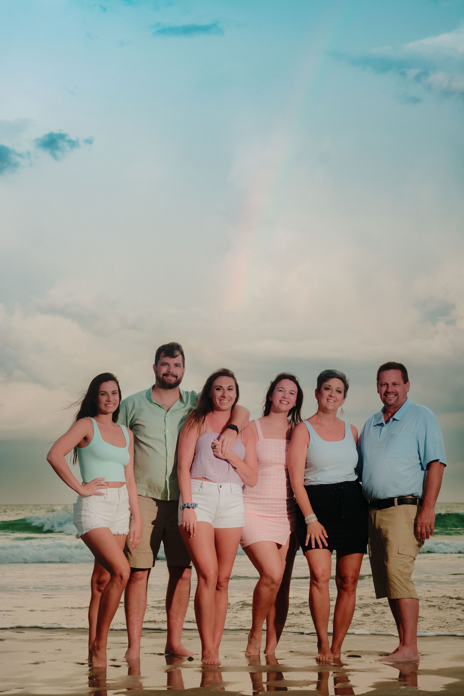 A group of people in light summer clothing pose together on a beach at sunset with turquoise sky and ocean waves.