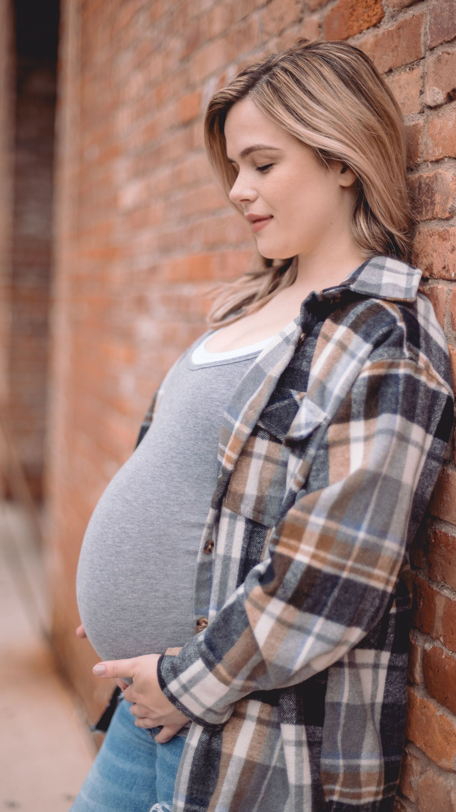 Expectant mother in plaid flannel shirt and jeans stands against brick wall while cradling her baby bump.
