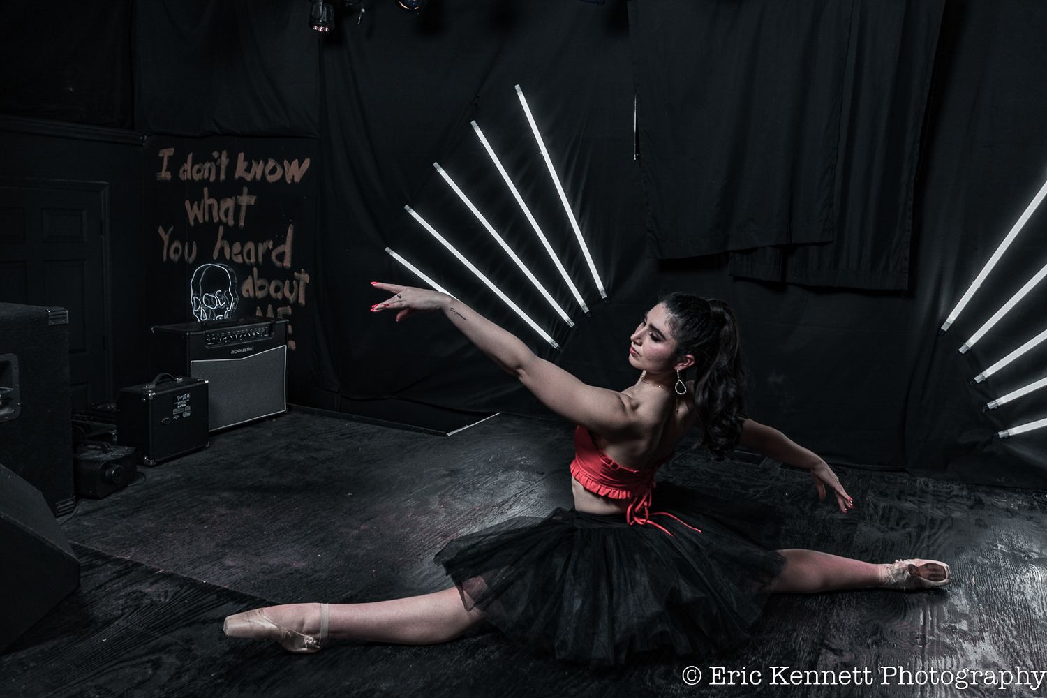 Dancer in red top and black tutu performs dramatic split pose against dark background with artistic white light rays.