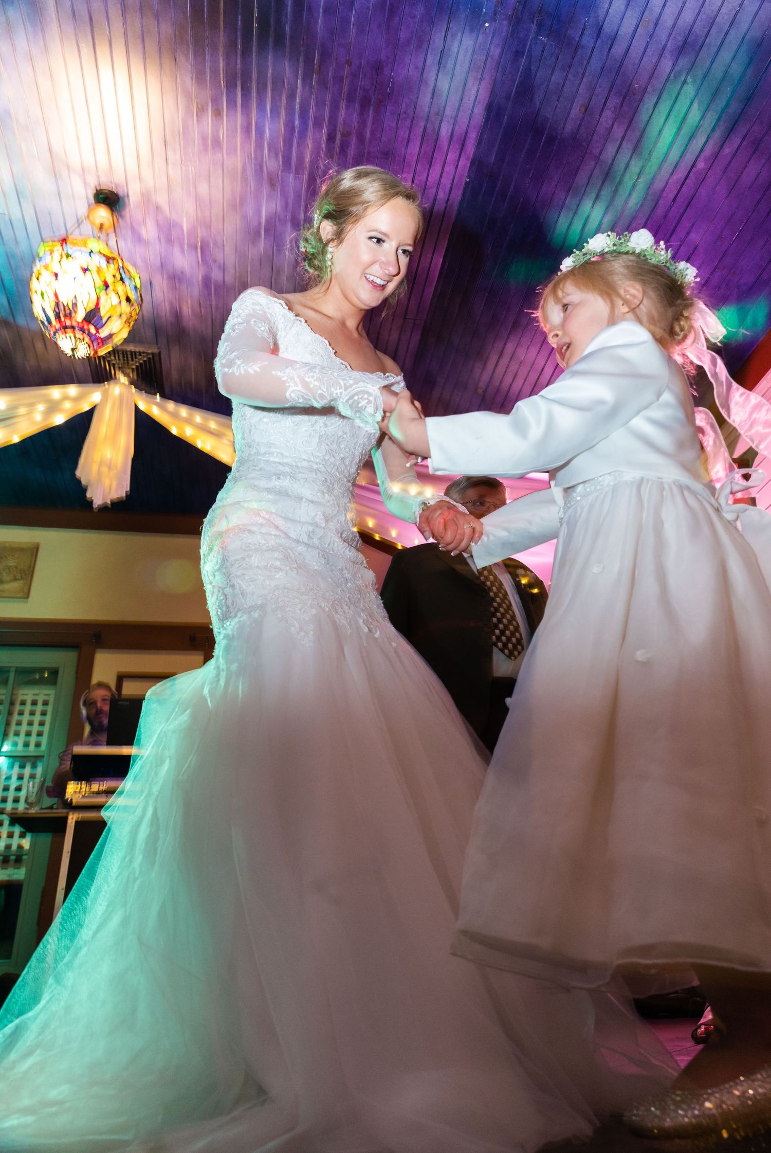 Two people in white wedding dresses dance together on a colorful illuminated dance floor during a celebration.