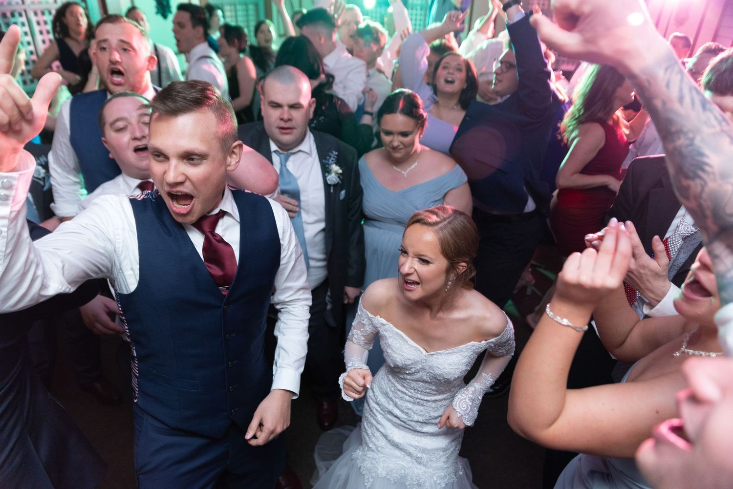 Wedding guests dancing and celebrating on a crowded dance floor with colorful lighting and festive atmosphere.