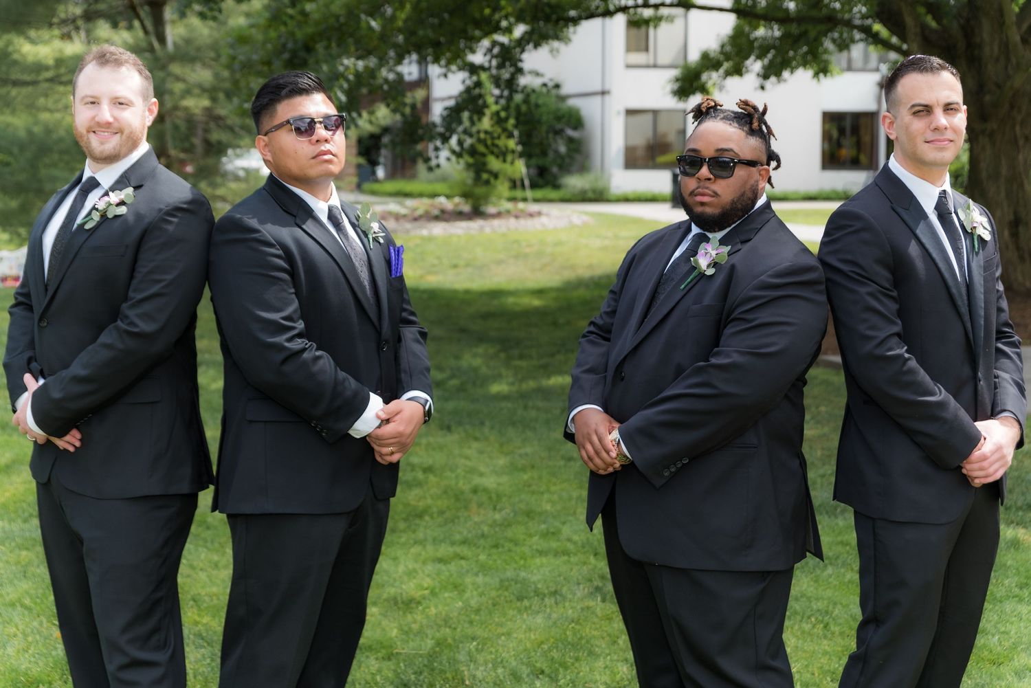 Four groomsmen in black suits and sunglasses pose together on a sunny lawn for a wedding photo.
