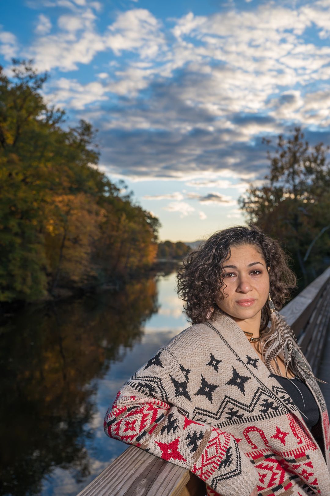 Person wearing a patterned sweater poses on a wooden bridge over a tranquil river during autumn sunset.