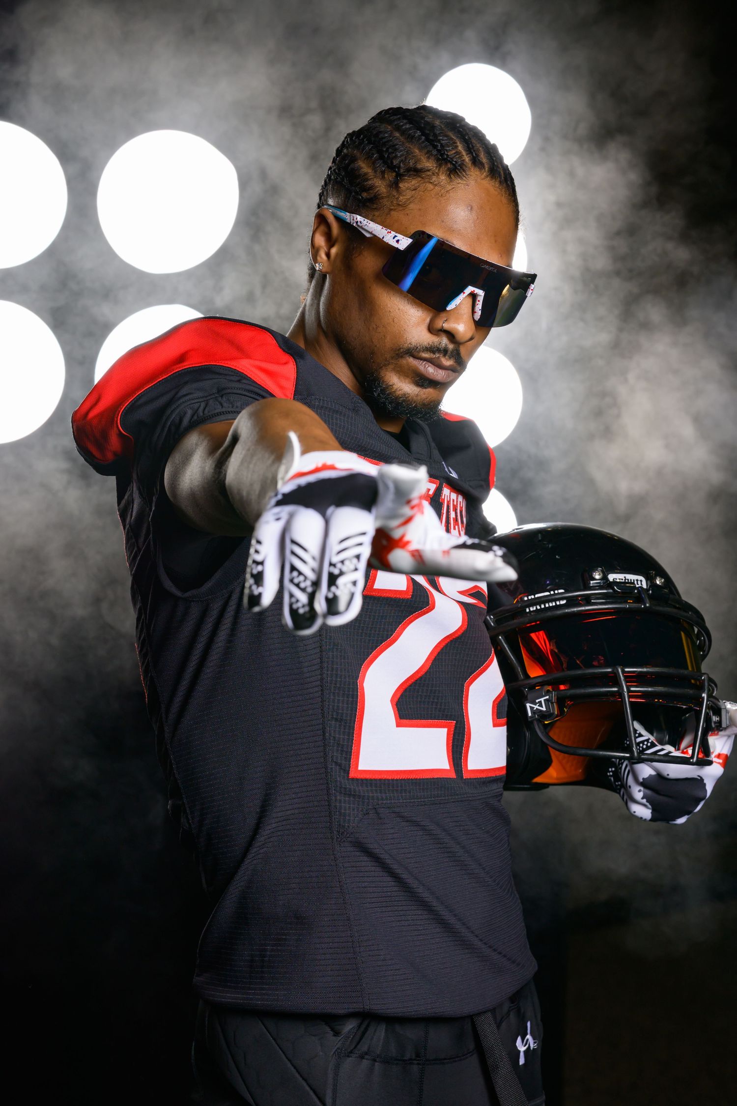 Football player in black jersey with number 22 poses in sunglasses and red hoodie against dramatic backlit backdrop.
