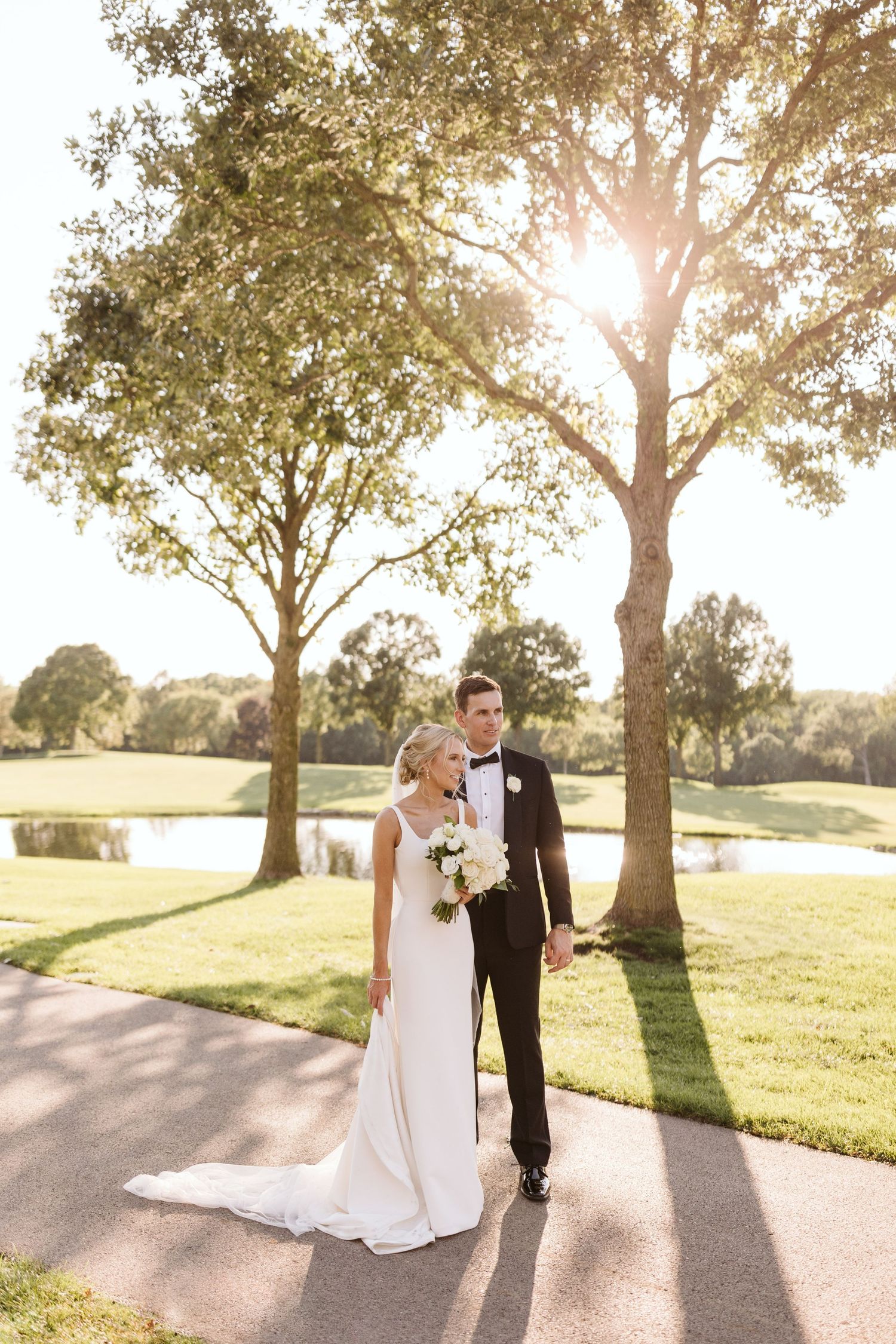 Wedding couple walks along a sunlit path on a golf course as golden sunlight streams through trees at sunset.