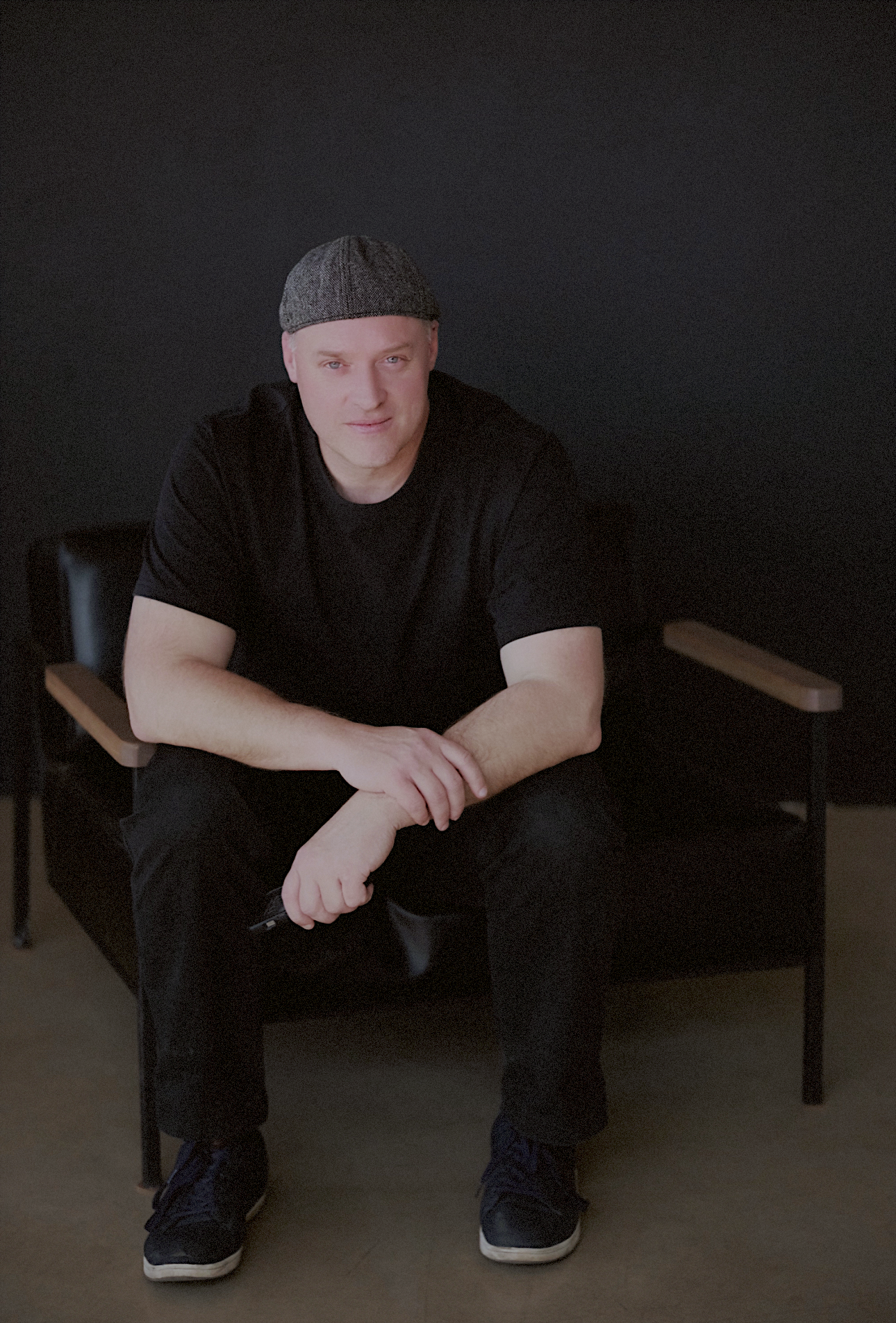 Man in black outfit and gray flat cap sitting relaxed on a leather chair against dark background.