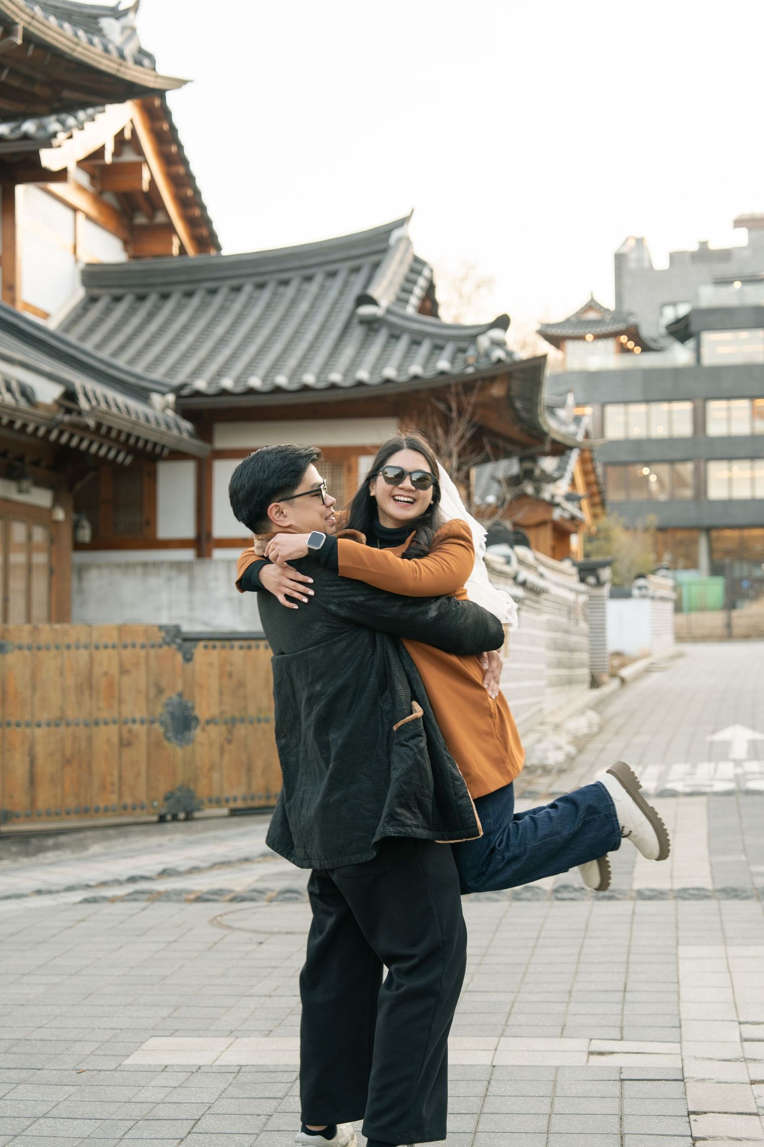 Two people embrace and laugh joyfully outside a traditional Korean hanok building on a city street.