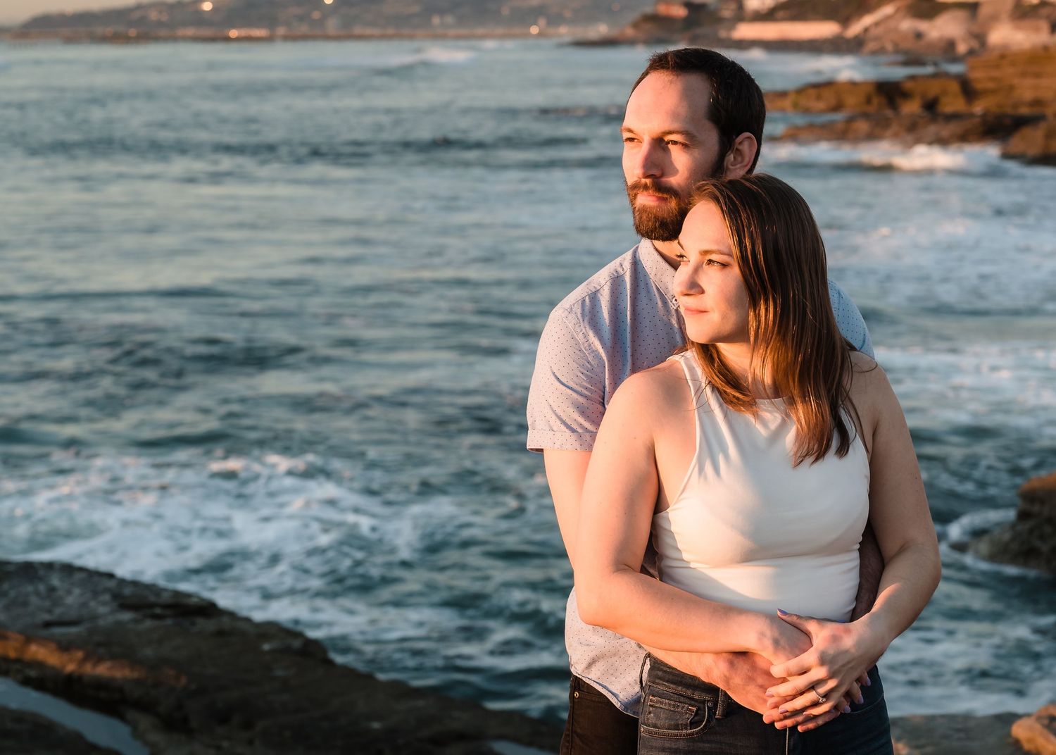 A romantic couple shares a tender moment on a rocky beach at sunset with ocean waves crashing in the background.
