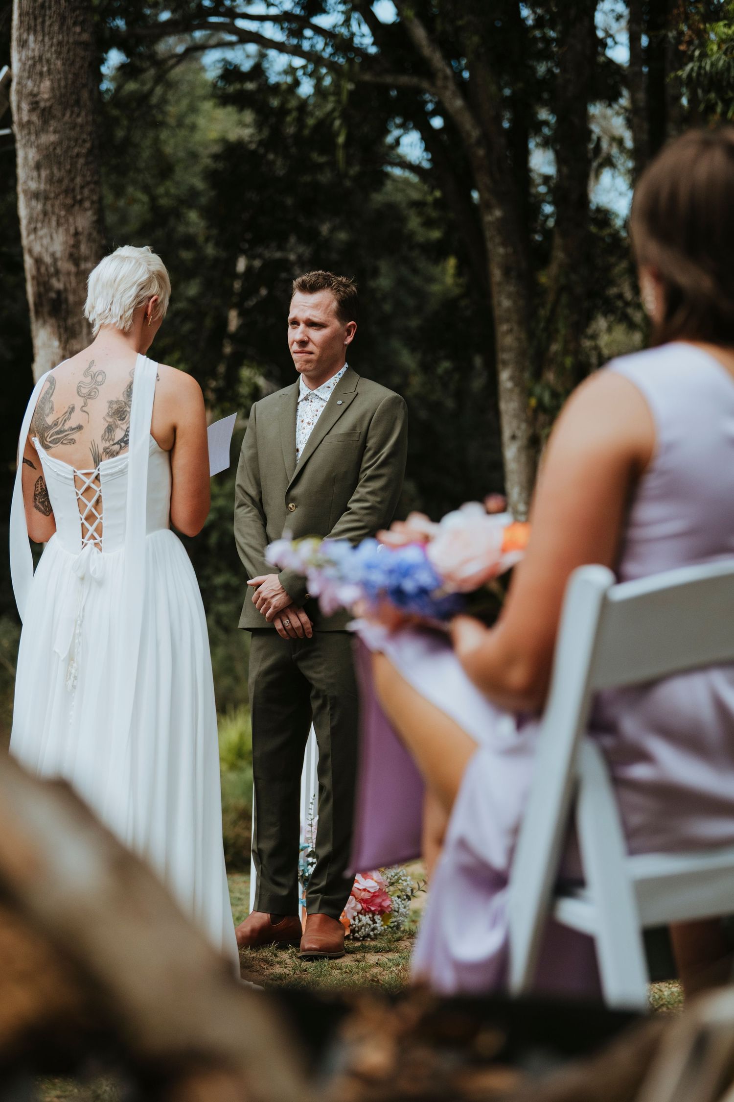 Wedding ceremony guests seated in white chairs watch as couple exchanges vows under trees in outdoor setting.