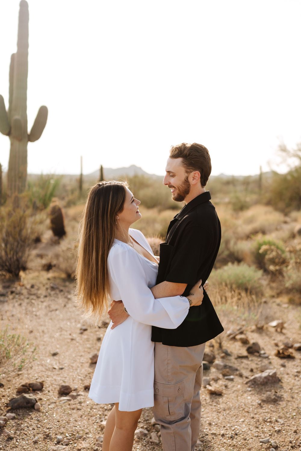 A couple shares a joyful moment together in a desert landscape with tall saguaro cacti in the background.