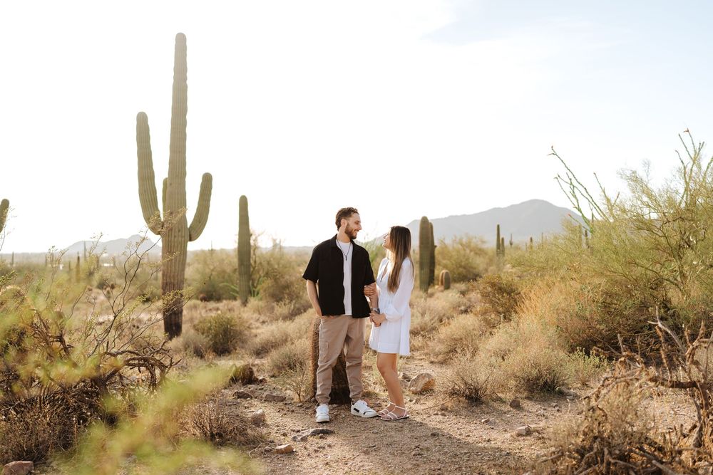 A couple stands together on a desert trail surrounded by towering saguaro cacti and desert landscape at sunset.