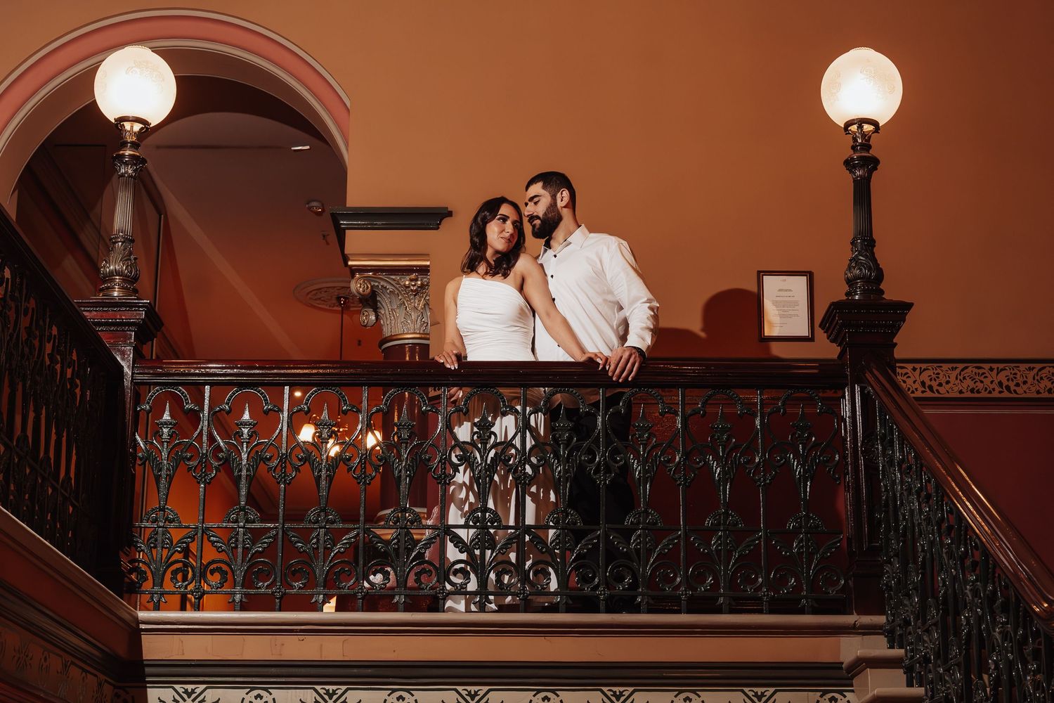 A couple in white outfits dances together on a red carpet in a dimly lit hallway with ornate lighting fixtures.
