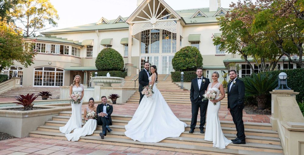 Wedding party poses on grand mansion steps with bride and groom at center.