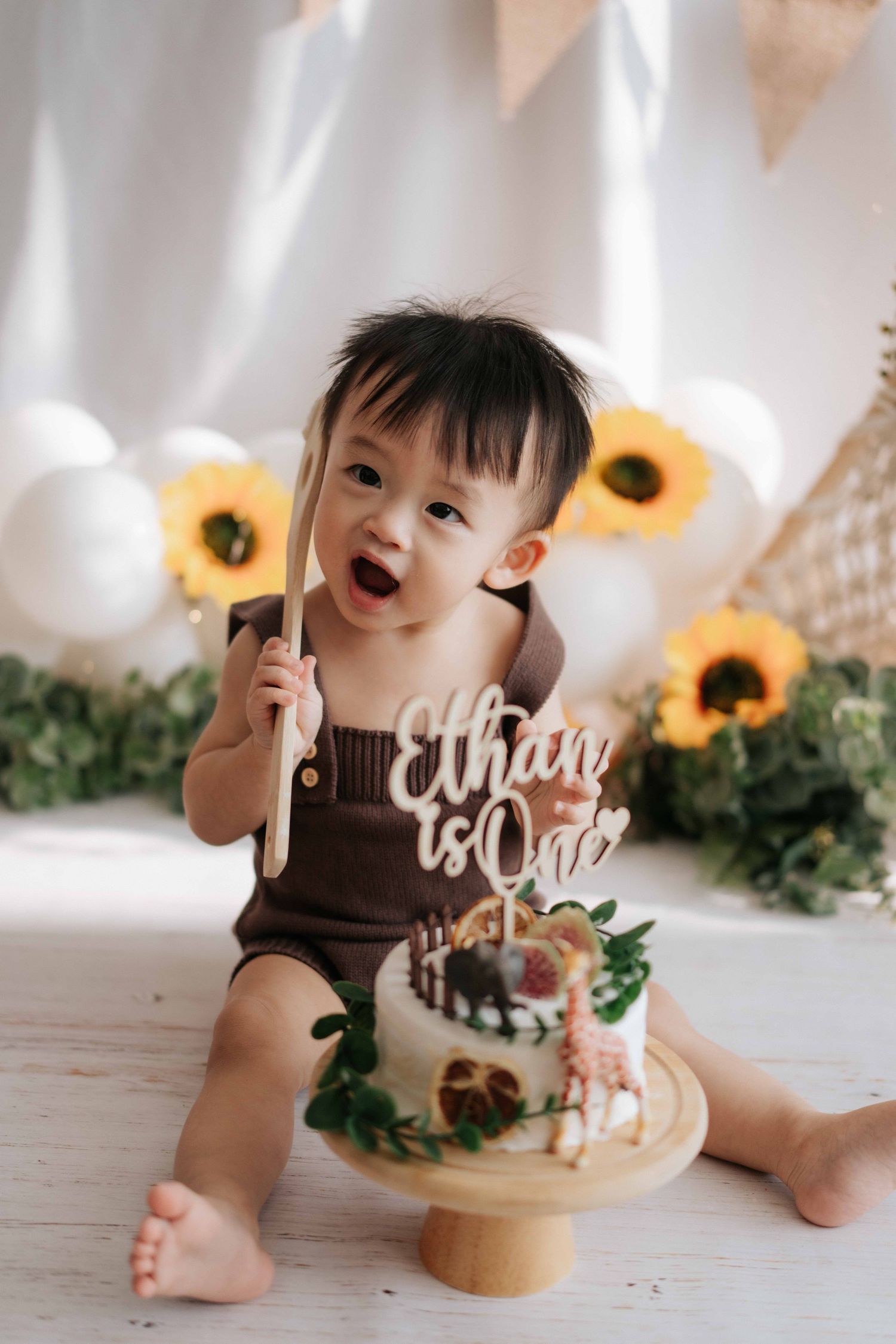 A baby in a brown romper sits next to a decorated cake with sunflowers and white lettering for a milestone celebration.