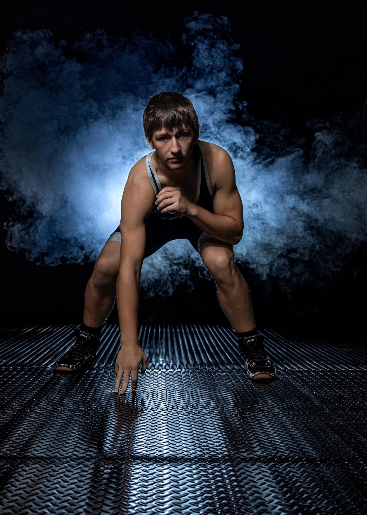 Athletic figure in dramatic low-angle pose on metallic floor with moody blue smoke effects in dark studio setting.