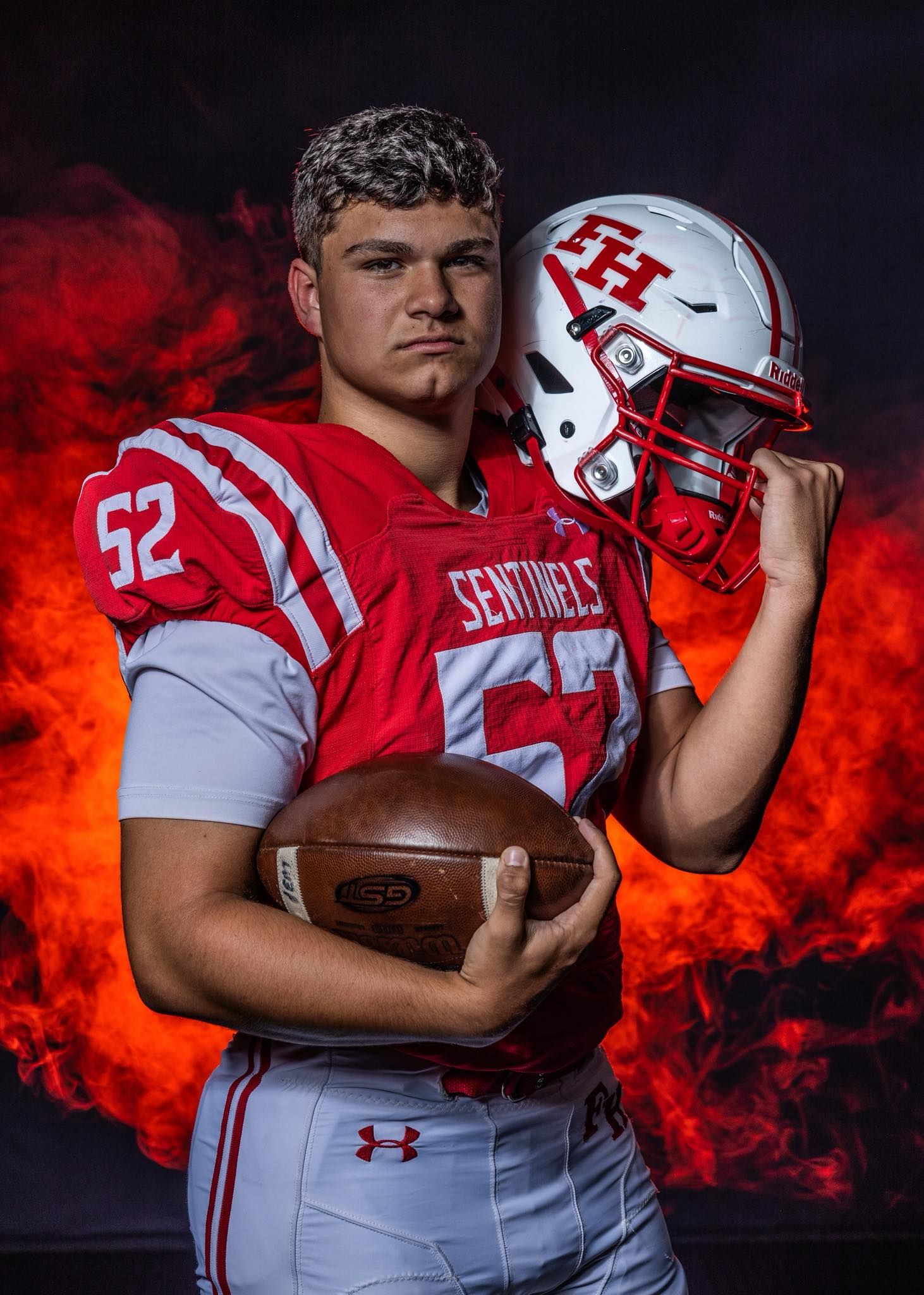 High school football player in red and white uniform holds football against dramatic fiery background.