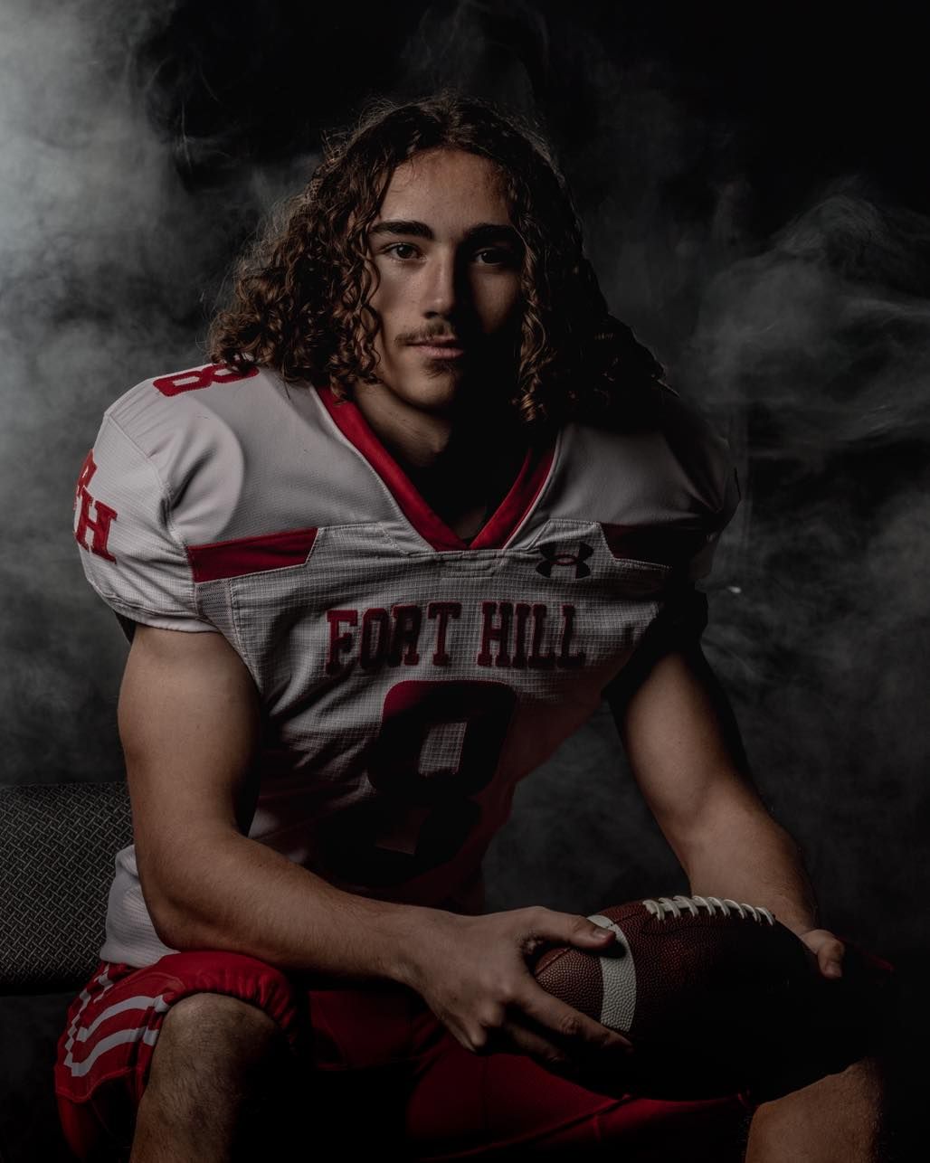 Dramatic sports portrait of a football player in white and red uniform holding ball against dark smoky background.