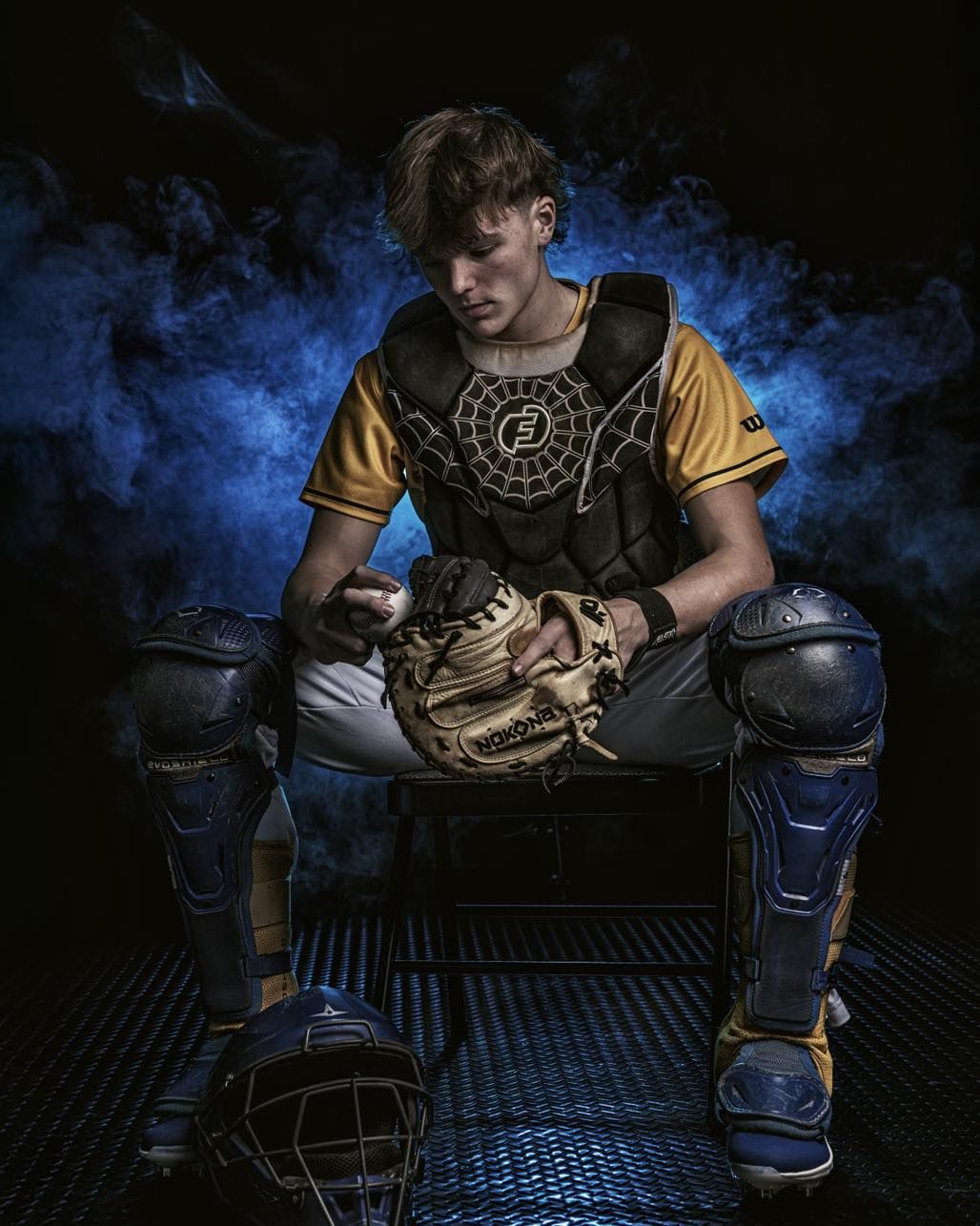 A baseball catcher in protective gear crouches in a dramatic low-light setting with blue and black tones.