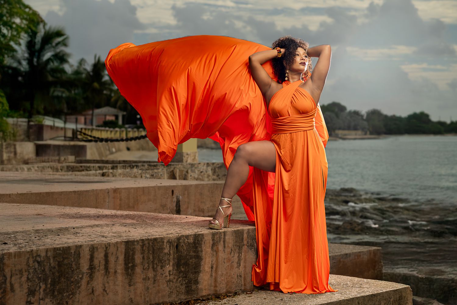 A person in a flowing orange dress poses gracefully along a waterfront stone wall at sunset.