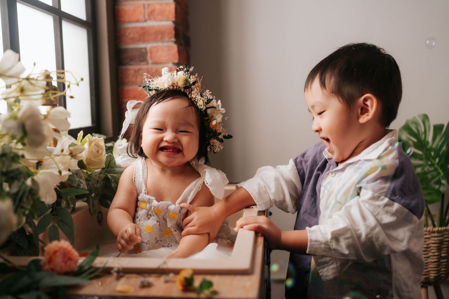 Baby in floral outfit plays near piano with white balloons and delicate floral arrangements in dreamy studio setting.